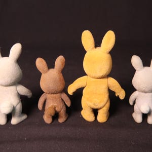 Może przedstawiać: Cztery małe, flokowane figurki kr&oacute;lik&oacute;w w r&oacute;żnych kolorach: jasnoszarym, brązowym, ż&oacute;łtym i szarym. Każdy ma wyprostowane uszy, mały ogon i wyciągnięte ramiona. Kr&oacute;liki stoją prosto.