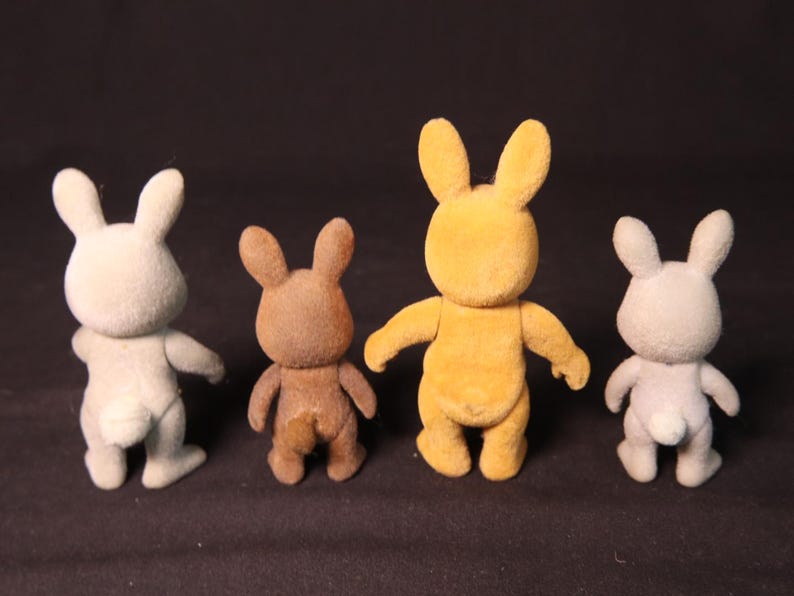 Może przedstawiać: Cztery małe, flokowane figurki kr&oacute;lik&oacute;w w r&oacute;żnych kolorach: jasnoszarym, brązowym, ż&oacute;łtym i szarym. Każdy ma wyprostowane uszy, mały ogon i wyciągnięte ramiona. Kr&oacute;liki stoją prosto.