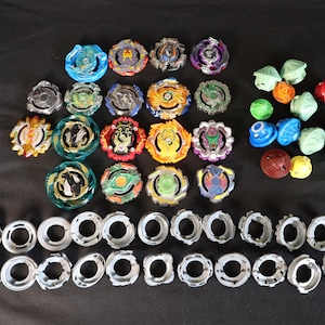 May include: A collection of colorful spinning tops and related components, including various tops in blue, green, orange, yellow, and purple. Also includes silver-colored rings and other accessories. The items are arranged on a black surface.