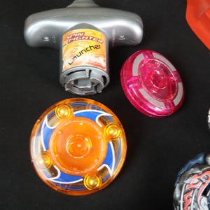 May include: A collection of colorful spinning tops and launcher toys. The tops feature orange, blue, and silver designs. The launcher is silver and has the words "Spin Fighter Launcher" printed on it. A red plastic piece is also visible.