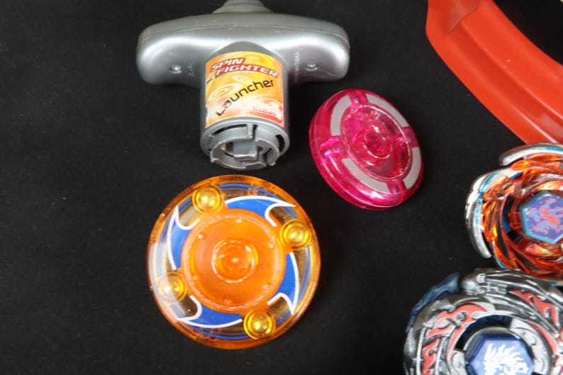May include: A collection of colorful spinning tops and launcher toys. The tops feature orange, blue, and silver designs. The launcher is silver and has the words "Spin Fighter Launcher" printed on it. A red plastic piece is also visible.