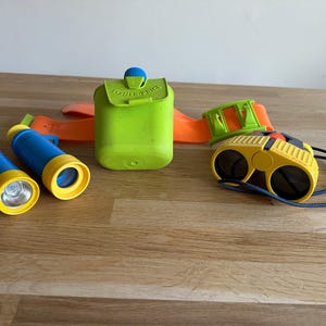 Op de afbeelding: Een kinderspeelset met een groene draagtas, een oranje riem, gele verrekijkers en twee blauw-gele zaklampen. De set is ontworpen voor fantasierijk spel en buitenavonturen.