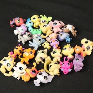 Puede incluir: Una colección de pequeñas figuras de animales de plástico de colores, que incluyen gatos, perros y pájaros. Los juguetes son en tonos de amarillo, rosa, azul y morado, con ojos grandes y expresivos.