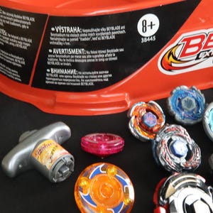 May include: A collection of colorful Beyblade spinning tops and a launcher, with an orange container in the background. The tops feature various designs and colors, including blue, orange, and silver. The image also shows the text "BEYBLADE EXTREME TOP SYSTEM RIP GRIND BEYSTADIUM".