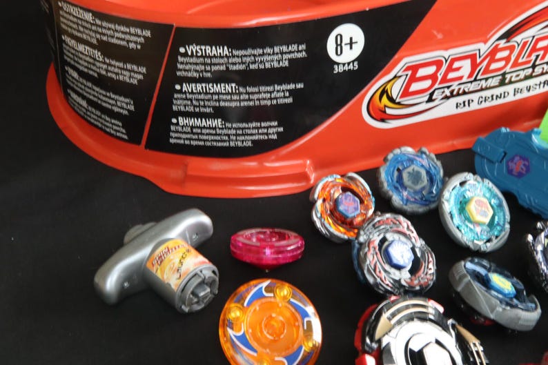 May include: A collection of colorful Beyblade spinning tops and a launcher, with an orange container in the background. The tops feature various designs and colors, including blue, orange, and silver. The image also shows the text "BEYBLADE EXTREME TOP SYSTEM RIP GRIND BEYSTADIUM".