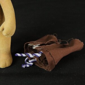 May include: A tan flocked toy figure standing next to a pair of brown fabric shorts. The shorts feature a small white and purple twisted cord drawstring at the waist, with visible stitching along the seams and a simple, unlined construction.