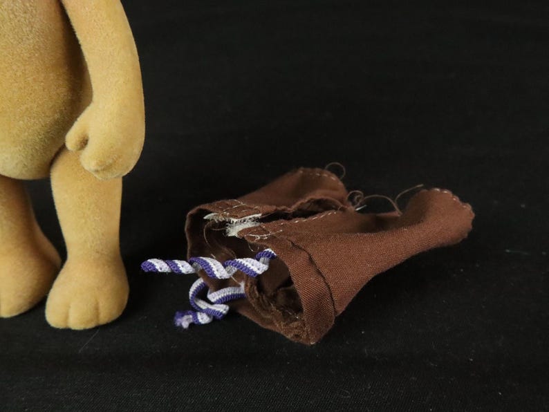 May include: A tan flocked toy figure standing next to a pair of brown fabric shorts. The shorts feature a small white and purple twisted cord drawstring at the waist, with visible stitching along the seams and a simple, unlined construction.