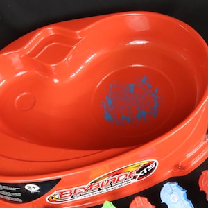 May include: An orange plastic Beyblade XTS Extreme Top System arena. The arena has a circular design with a recessed area and a blue graphic in the center. Accessories, including launchers, are visible.