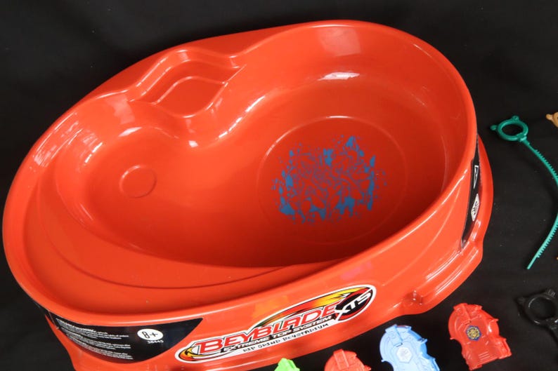 May include: An orange plastic Beyblade XTS Extreme Top System arena. The arena has a circular design with a recessed area and a blue graphic in the center. Accessories, including launchers, are visible.