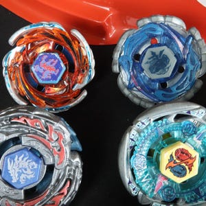 May include: A collection of colorful spinning tops in various designs. The tops feature metallic silver, blue, orange, and teal accents. Each top has a central emblem with detailed graphics. A red plastic arena is visible in the background.