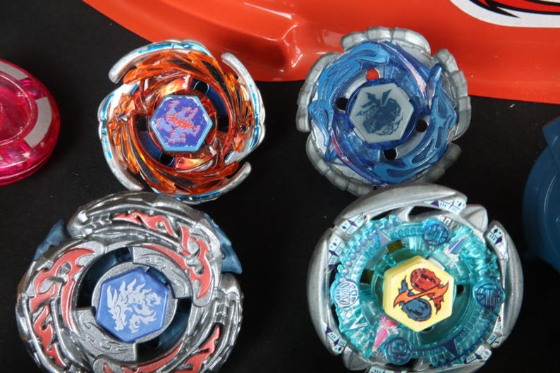 May include: A collection of colorful spinning tops in various designs. The tops feature metallic silver, blue, orange, and teal accents. Each top has a central emblem with detailed graphics. A red plastic arena is visible in the background.