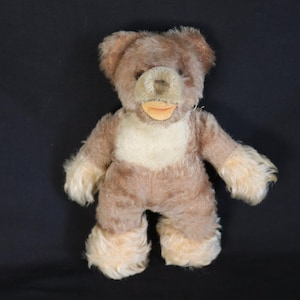 Puede incluir: Un oso de peluche vintage con cuerpo marrón claro, pecho crema y detalles color canela en la cabeza, patas y pies. El oso tiene boca cosida y hocico crema. El peluche está sobre un fondo oscuro.