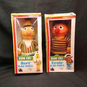 Op de afbeelding: Twee vintage Sesame Street lappenpoppen in originele verpakking. De linkerdoos bevat een gele Bert-pop met een gestreept shirt. De rechterdoos bevat een oranje Ernie-pop met een veelkleurige gestreepte trui. Beide dozen tonen het Knickerbocker-logo.