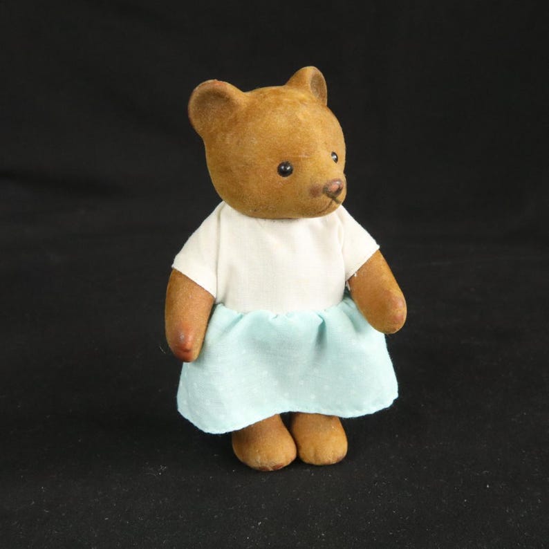 Peut inclure: Une petite figurine d'ours marron &agrave; la surface textur&eacute;e, portant un haut blanc &agrave; manches courtes et une jupe bleu clair avec un motif discret de pois blancs. L'ours a des yeux sombres, un petit nez et des oreilles arrondies, debout.