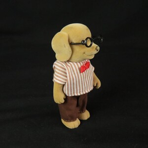 May include: A tan dog figurine with a fuzzy texture, wearing black round-rimmed glasses, a white and brown vertical striped short-sleeve shirt, a small red bow tie at the collar, and solid brown trousers, standing against a plain black background.