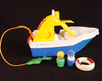 Puede incluir: Un barco de juguete vintage con casco azul, cubierta blanca y superestructura amarilla. Incluye dos pequeñas figuras, un caimán verde y un salvavidas blanco con rayas rojas. Una cuerda naranja está unida al caimán.