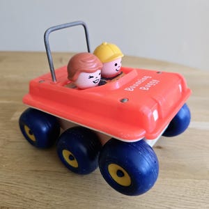 Puede incluir: Un coche de juguete rojo y blanco vintage con ruedas azules y detalles amarillos. El coche de juguete tiene dos pequeñas figuras de plástico en el interior. Las palabras "Bouncing Buggy" están impresas en el lateral. Una barra de metal sirve como asa.