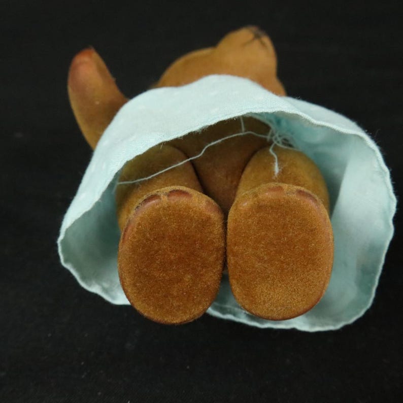 Peut inclure: Une petite figurine d'ours en peluche floqu&eacute; marron avec une texture duveteuse, portant une jupe bleu clair avec un motif discret &agrave; pois blancs. L'ours est vu d'en haut, r&eacute;v&eacute;lant ses pieds arrondis et le tissu de la jupe.