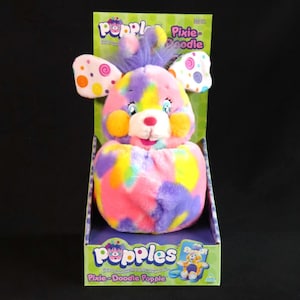 Könnte beinhalten: Ein farbenfrohes Pixie-Doodle Popple Plüschtier in seiner Originalverpackung. Das Plüschtier hat eine Mischung aus rosa, lila, gelbem und blauem Fell. Die Box ist grün mit den Wörtern "Popples" und "Pixie-Doodle Popple" bedruckt.