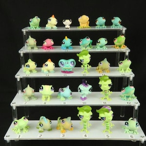 May include: A collection of twenty-nine small, stylised plastic frog and lizard figurines arranged on a five-tiered white acrylic display stand. Each figurine features large eyes and is numbered with a small black digit on the shelf surface.