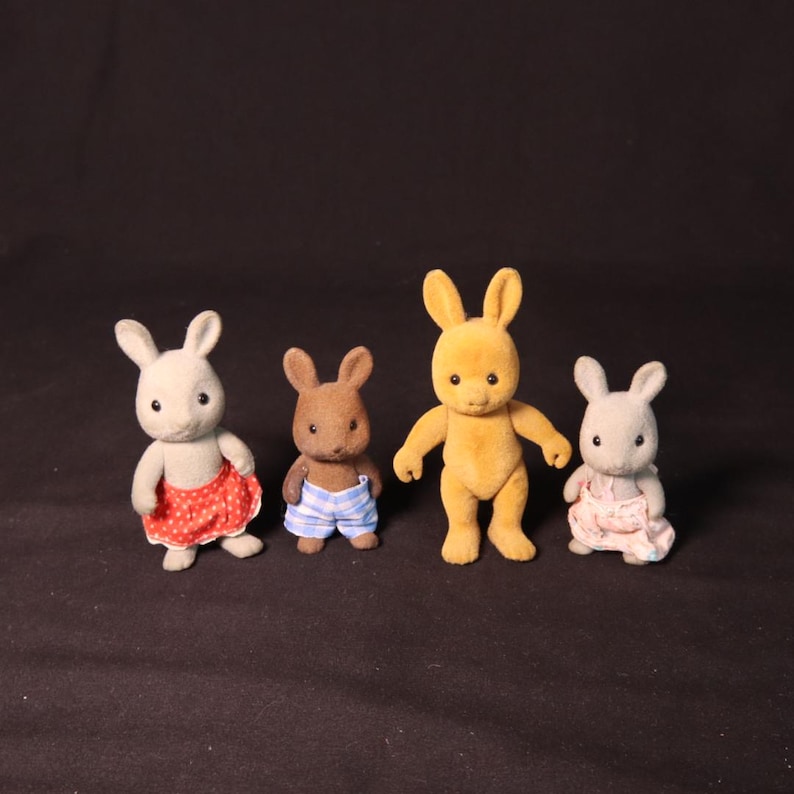 Może przedstawiać: Cztery figurki zabawek kr&oacute;lik&oacute;w. Jedna jest szara z czerwoną sp&oacute;dnicą w kropki, druga jest brązowa z niebiesko-białymi szortami w paski, trzecia jest ż&oacute;łta, a ostatnia szara z r&oacute;żową sukienką. Wszystkie mają długie uszy.