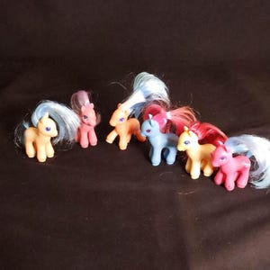以下が含まれることがあります： 6匹の小さなカラフルなおもちゃのポニーのコレクション。ポニーは黄色、ピンク、オレンジ、青など様々な色があります。各ポニーは異なる色のたてがみと尾を持っています。ポニーは暗い表面に立っています。