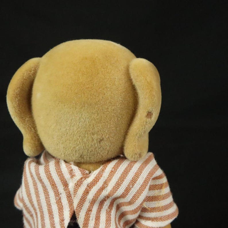 May include: A small, tan, flocked animal figurine viewed from the back, featuring rounded ears and a textured, fuzzy surface. The figurine is wearing a shirt made of white fabric with thin, vertical, light brown stripes that cross at the neck.