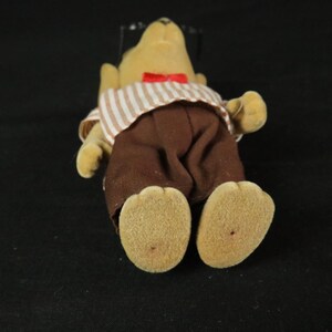 May include: A flocked tan animal figurine wearing a white and brown striped shirt with a red bow tie and solid brown trousers. The figure has a rounded snout, small ears, and textured feet, presented in a standing position against a dark background.