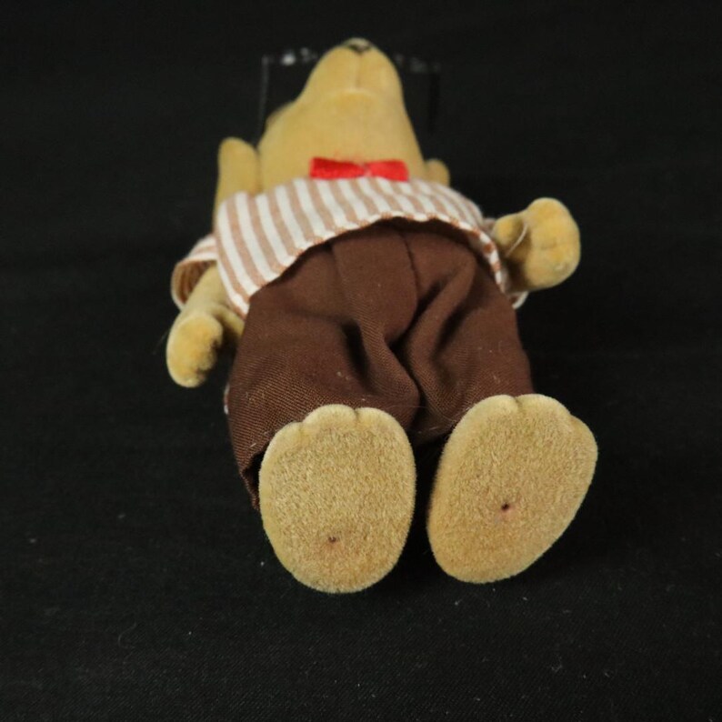 May include: A flocked tan animal figurine wearing a white and brown striped shirt with a red bow tie and solid brown trousers. The figure has a rounded snout, small ears, and textured feet, presented in a standing position against a dark background.