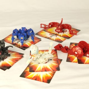 Peut inclure: Plusieurs figurines de jouets colorées, dont une créature bleue en forme d'araignée, un dragon noir et des figurines rouges segmentées, sont présentées sur des cartes au design flamboyant. Les jouets sont posés sur un fond blanc.