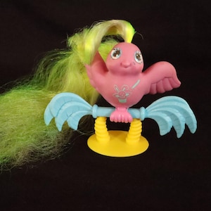 Op de afbeelding: Een vintage roze vogelbeeldje met blauwe vleugels en een gele basis. De vogel heeft een lange, vloeiende groen en gele staart. Een klein bloemenontwerp staat op de borst van de vogel. Dit verzamelbare speelgoed is een nostalgisch item.