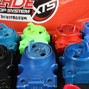May include: A collection of colorful toy tops in various shades of blue, green, red, and black. The tops have a unique design with a central spinning mechanism and a hook-like feature. The background includes text that reads "Beyblade Extreme Top System".