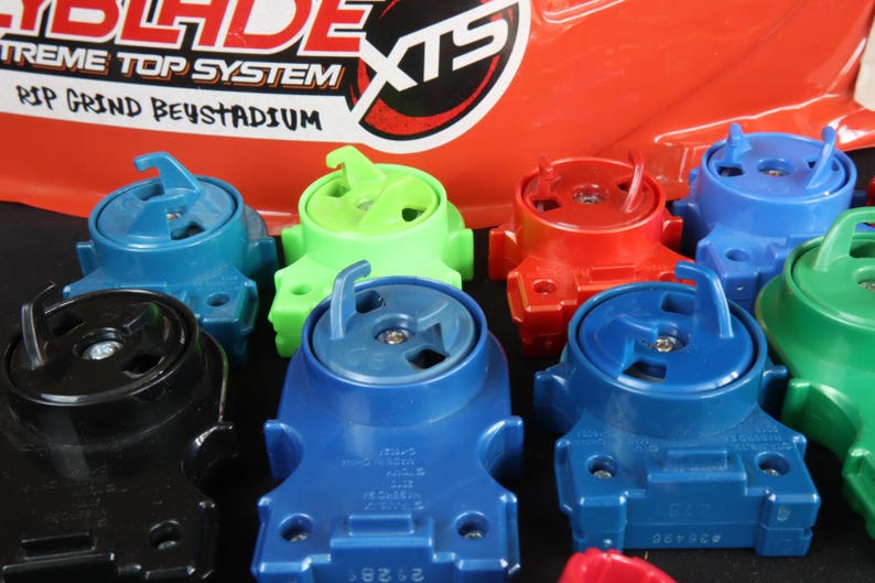 May include: A collection of colorful toy tops in various shades of blue, green, red, and black. The tops have a unique design with a central spinning mechanism and a hook-like feature. The background includes text that reads "Beyblade Extreme Top System".