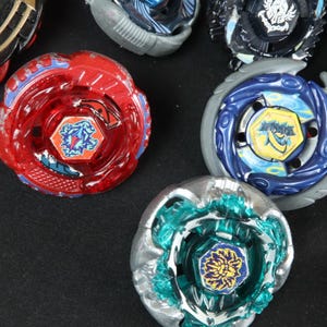 May include: A collection of colorful spinning tops, including red, blue, and teal designs. Each top features intricate details and a central emblem. The toys are arranged on a dark surface, showcasing their vibrant colors and unique patterns.