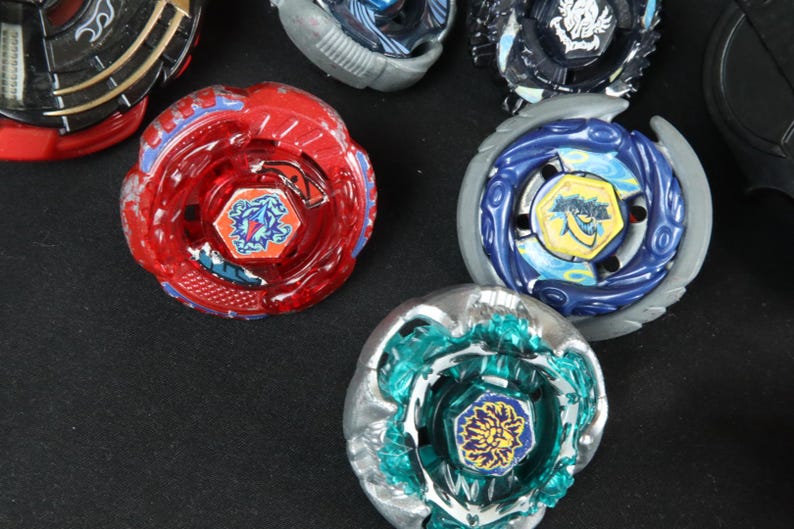 May include: A collection of colorful spinning tops, including red, blue, and teal designs. Each top features intricate details and a central emblem. The toys are arranged on a dark surface, showcasing their vibrant colors and unique patterns.
