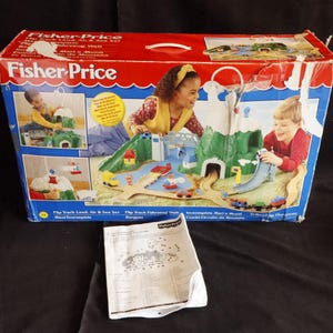 以下が含まれることがあります： Fisher-Price Flip Track Land、Air & Sea Setのおもちゃ箱。箱は赤と青で、子供たちが玩具セットで遊んでいる画像が描かれています。玩具セットには、山、トラック、乗り物、橋が含まれています。