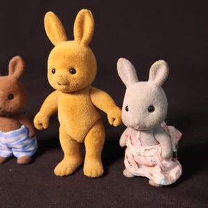 Może przedstawiać: Trzy figurki zabawek kr&oacute;lik&oacute;w. Centralny kr&oacute;lik jest w kolorze złotoż&oacute;łtym, w pozycji stojącej. Po lewej stronie znajduje się brązowy kr&oacute;lik w niebiesko-białych szortach w paski. Po prawej stronie szary kr&oacute;lik w r&oacute;żowej sukience.