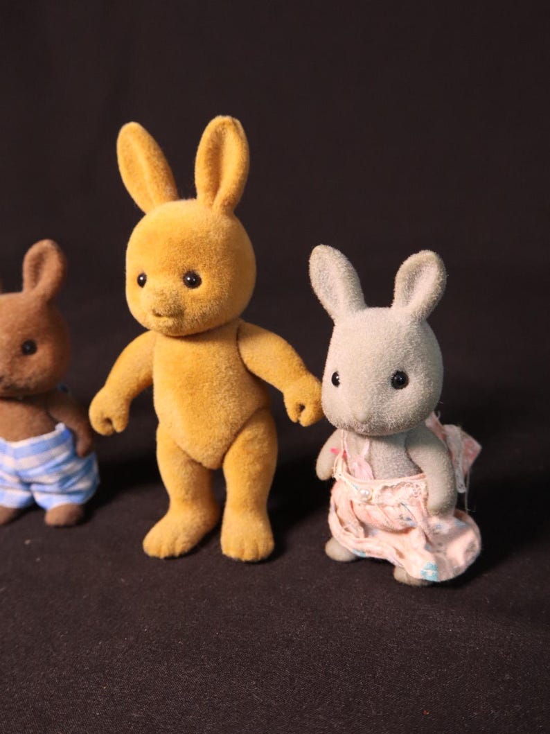 Może przedstawiać: Trzy figurki zabawek kr&oacute;lik&oacute;w. Centralny kr&oacute;lik jest w kolorze złotoż&oacute;łtym, w pozycji stojącej. Po lewej stronie znajduje się brązowy kr&oacute;lik w niebiesko-białych szortach w paski. Po prawej stronie szary kr&oacute;lik w r&oacute;żowej sukience.