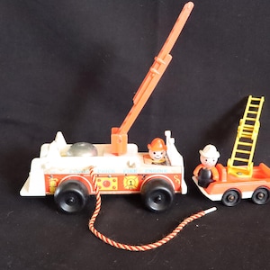 Könnte beinhalten: Ein Vintage-Feuerwehrauto-Spielzeugset von Fisher-Price. Das Set enthält ein weißes und rotes Feuerwehrauto mit einer Leiter, ein kleines orangefarbenes Feuerwehrauto mit einer gelben Leiter und zwei Feuerwehrmannfiguren. Das Feuerwehrauto hat schwarze Räder und eine rot-weiße Schnur.