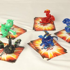 Peut inclure: Cinq figurines Bakugan colorées, dont vertes, rouges, bleues et noires, sont présentées sur des cartes de jeu au design flamboyant. Les jouets sont dans diverses poses, mettant en valeur leurs designs et caractéristiques uniques.