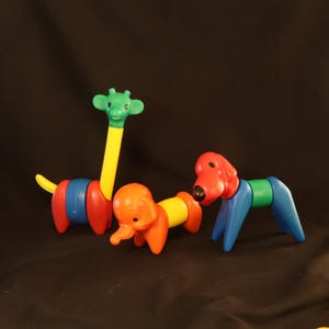 Può includere: Tre giocattoli di animali in plastica colorati: una giraffa, un elefante e un cane. La giraffa ha la testa verde e il collo giallo, l'elefante è arancione e il cane è rosso. Ogni giocattolo è composto da più pezzi.
