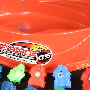 May include: An orange Beyblade Extreme Top System stadium with several colorful Beyblade tops. The stadium has a large, circular design with the Beyblade logo. The tops are red, blue, and green.