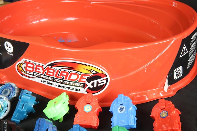 May include: An orange Beyblade Extreme Top System stadium with several colorful Beyblade tops. The stadium has a large, circular design with the Beyblade logo. The tops are red, blue, and green.