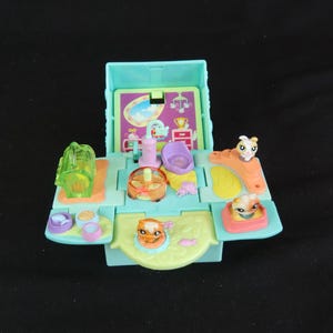 May include: A vintage, open playset in shades of teal, orange, and yellow. The playset features miniature animal figures, a small house, and various accessories. The design includes a small pool, a bed, and food bowls. The playset is designed for imaginative play.