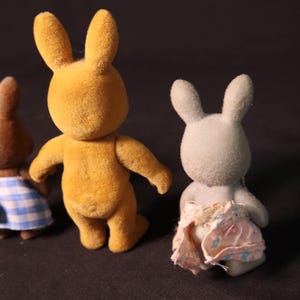 Może przedstawiać: Trzy figurki zabawek kr&oacute;lik&oacute;w. Jedna jest złotoż&oacute;łta, druga brązowa ze sp&oacute;dnicą w niebiesko-białą kratę, a trzecia jasnoszara z r&oacute;żową pieluchą. Kr&oacute;liki mają długie uszy i stoją.