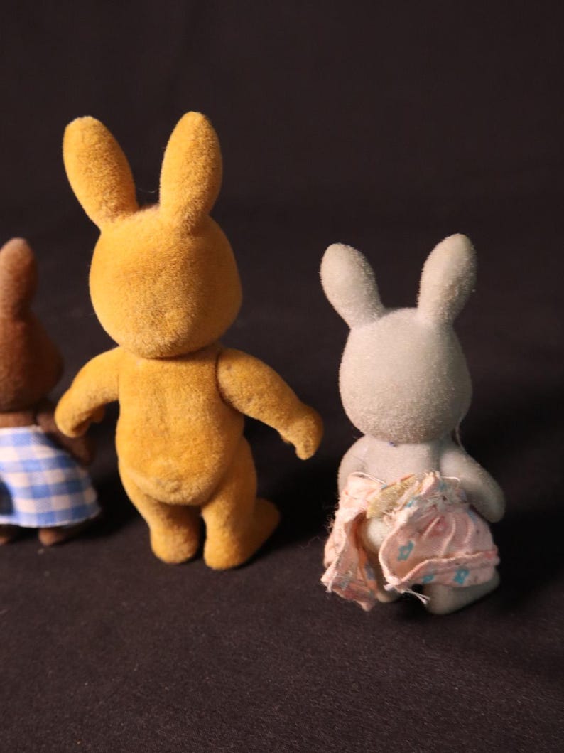 Może przedstawiać: Trzy figurki zabawek kr&oacute;lik&oacute;w. Jedna jest złotoż&oacute;łta, druga brązowa ze sp&oacute;dnicą w niebiesko-białą kratę, a trzecia jasnoszara z r&oacute;żową pieluchą. Kr&oacute;liki mają długie uszy i stoją.