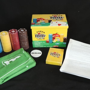 May include: A yellow box poker set with the M&M's logo, poker chips in blue, yellow, red, and brown, a green felt mat, a dealer button, and a deck of cards. The box features M&M's characters.