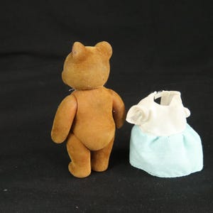 Peut inclure: Une figurine d'ours en peluche floqu&eacute; marron debout, plac&eacute;e &agrave; c&ocirc;t&eacute; d'une petite robe de poup&eacute;e. La robe pr&eacute;sente un corsage blanc &agrave; manches courtes et une jupe bleu clair avec un motif &agrave; pois blancs, con&ccedil;ue pour une figurine miniature.