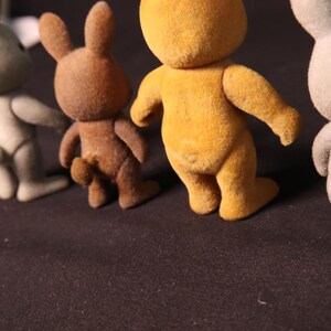 Może przedstawiać: Kolekcja małych, flokowanych figurek kr&oacute;lik&oacute;w w r&oacute;żnych kolorach, w tym szarym, brązowym i ż&oacute;łtym. Kr&oacute;liki stoją na ciemnej powierzchni, tyłem do widza. Figurki mają małe ogonki i wyprostowane uszy.