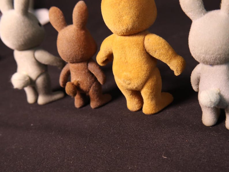 Może przedstawiać: Kolekcja małych, flokowanych figurek kr&oacute;lik&oacute;w w r&oacute;żnych kolorach, w tym szarym, brązowym i ż&oacute;łtym. Kr&oacute;liki stoją na ciemnej powierzchni, tyłem do widza. Figurki mają małe ogonki i wyprostowane uszy.